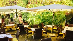 Un patio al aire libre con mesas y sillas, cubierto por sombrillas, rodeado de abundante vegetación.