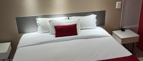 Une chambre d’hôtel avec un grand lit, du linge de lit blanc, un coussin décoratif rouge, une tête de lit, une table de chevet et une petite table de nuit.