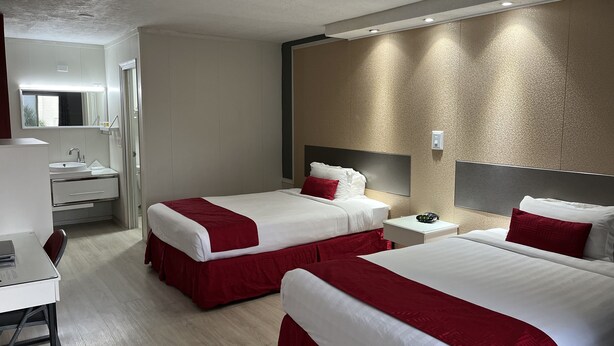 A hotel room with two beds, a desk, a chair, and a bathroom with a sink and mirror.