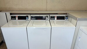 A row of three white commercial washers with control panels, located in a utility room with a tiled floor and a plain wall.