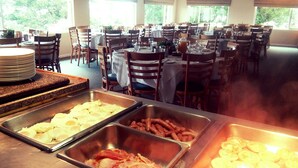 A buffet setup with various food items like eggs, bacon, and sausages. Tables are set with white tablecloths and surrounded by wooden chairs. - Carmel Guest Farm (Wilderness)