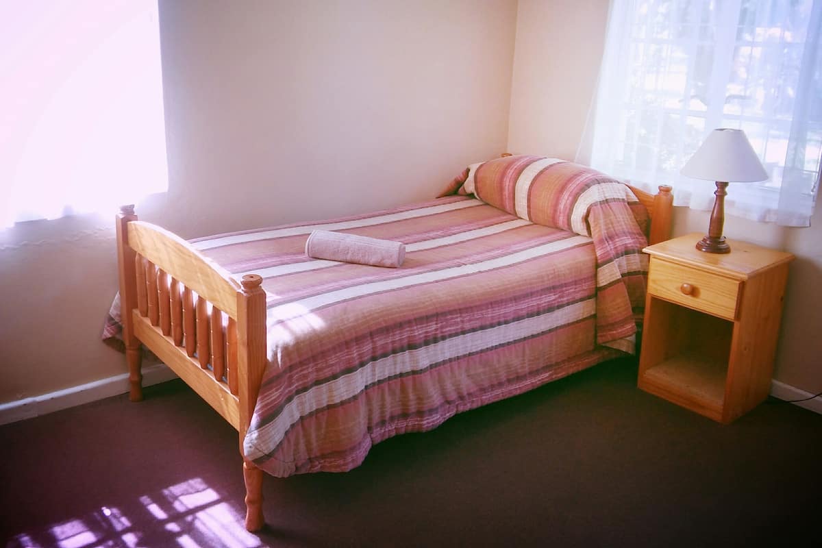 A single bed with a striped bedspread and a wooden headboard, a nightstand with a lamp, and a window with curtains.