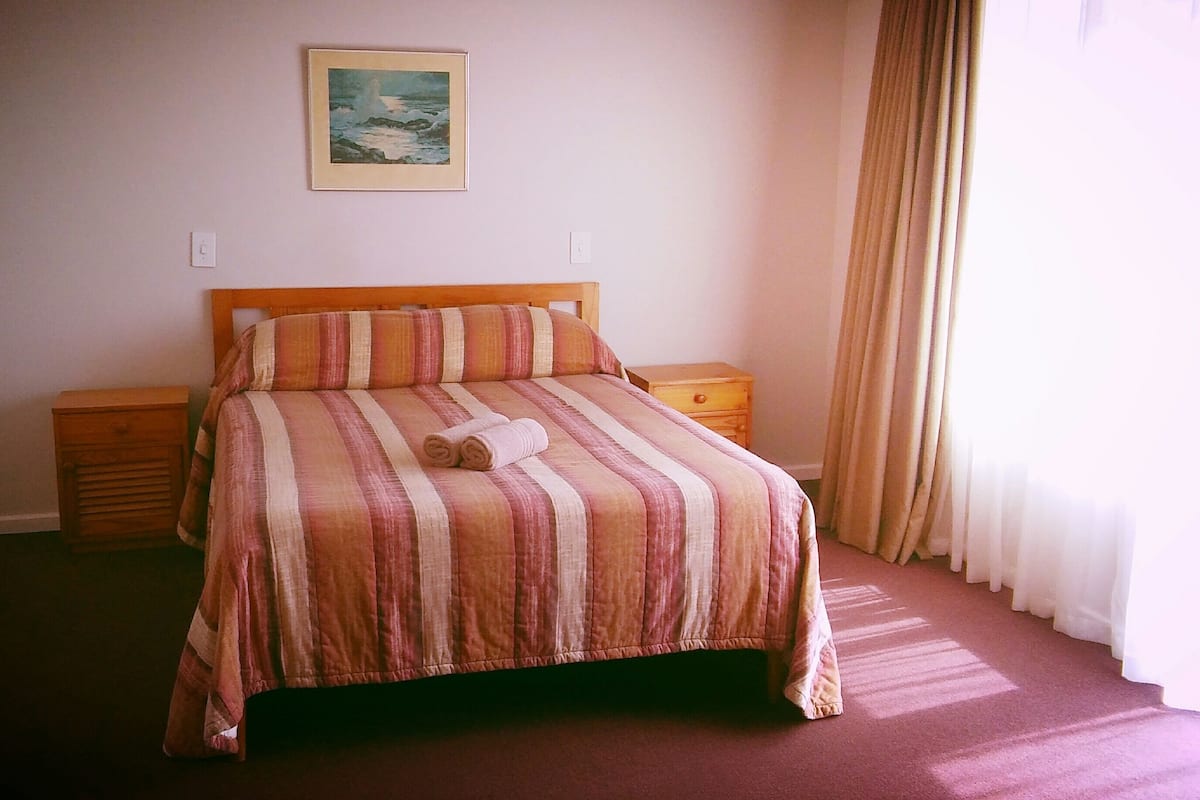 A neatly made bed with striped bedding, two bedside tables, a nightstand, a framed picture on the wall, and a window with curtains.