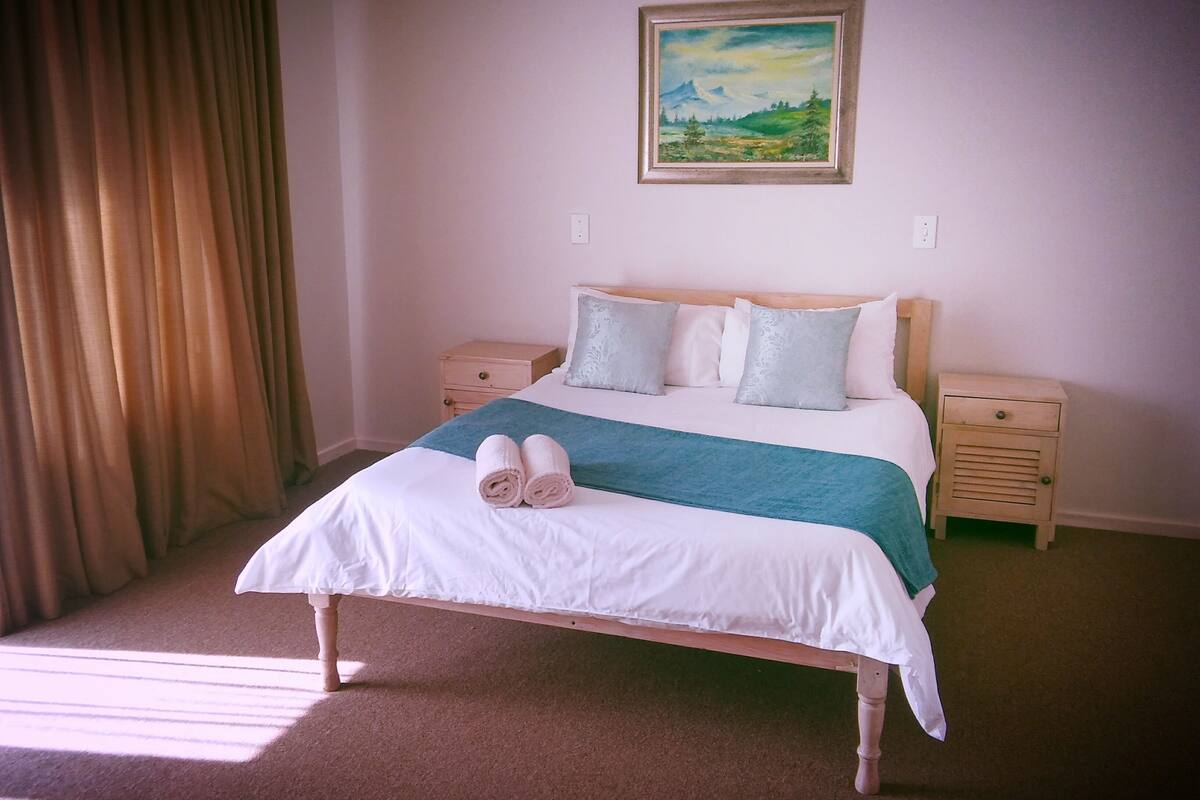 A neatly made bed with a white and green bedspread, two pillows, and rolled towels. There is a painting of a landscape on the wall above the bed.