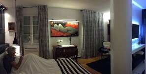 A bedroom with a bed, a bedside table with a lamp, a painting on the wall, and a window with curtains. - Le 61 Berthier (Paris)