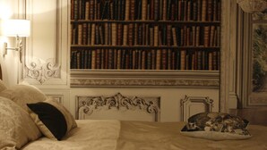 A bedroom with a large bookshelf filled with books, a bed with patterned bedding, and a decorative headboard. - Kathome (Bondues)