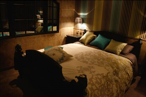 A dimly lit hotel room with a large bed, a nightstand, a lamp, and a window with blinds.