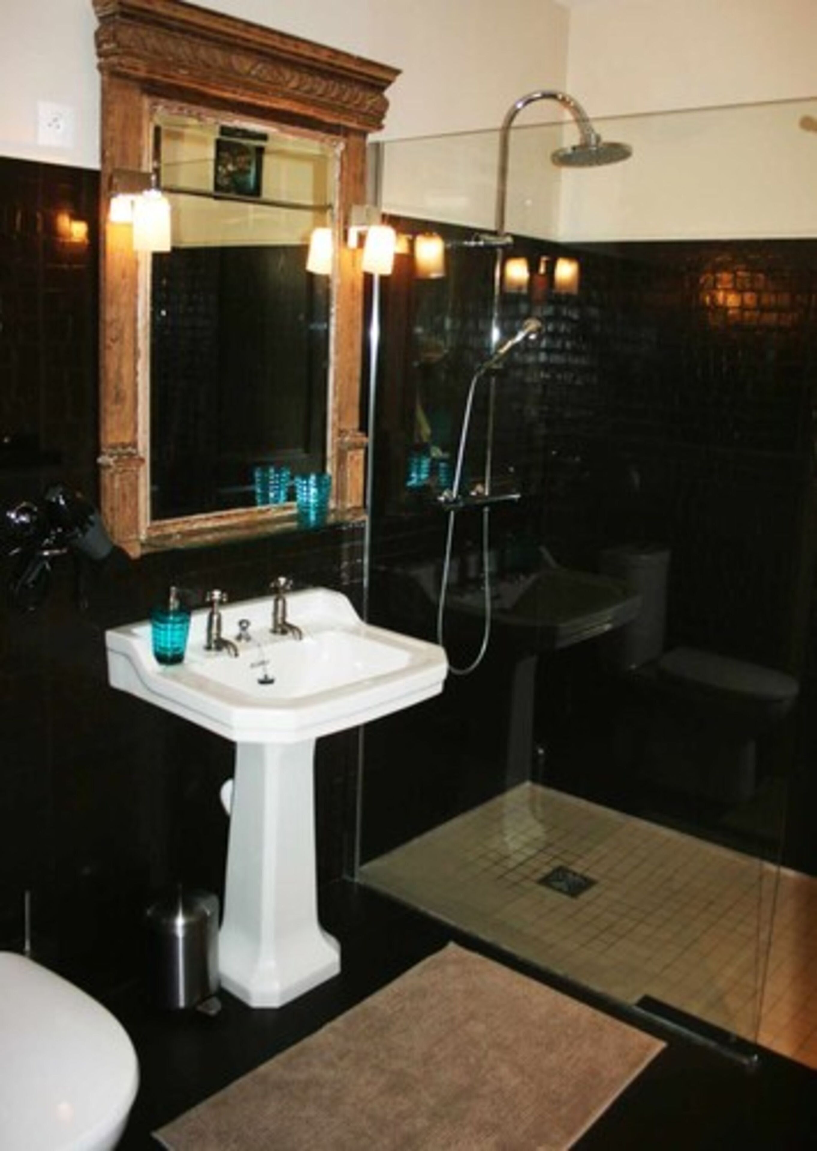 Een badkamer met een wit wastafeltje op een voet, een spiegel met houten lijst, een douche met glazen wanden, en een toilet.