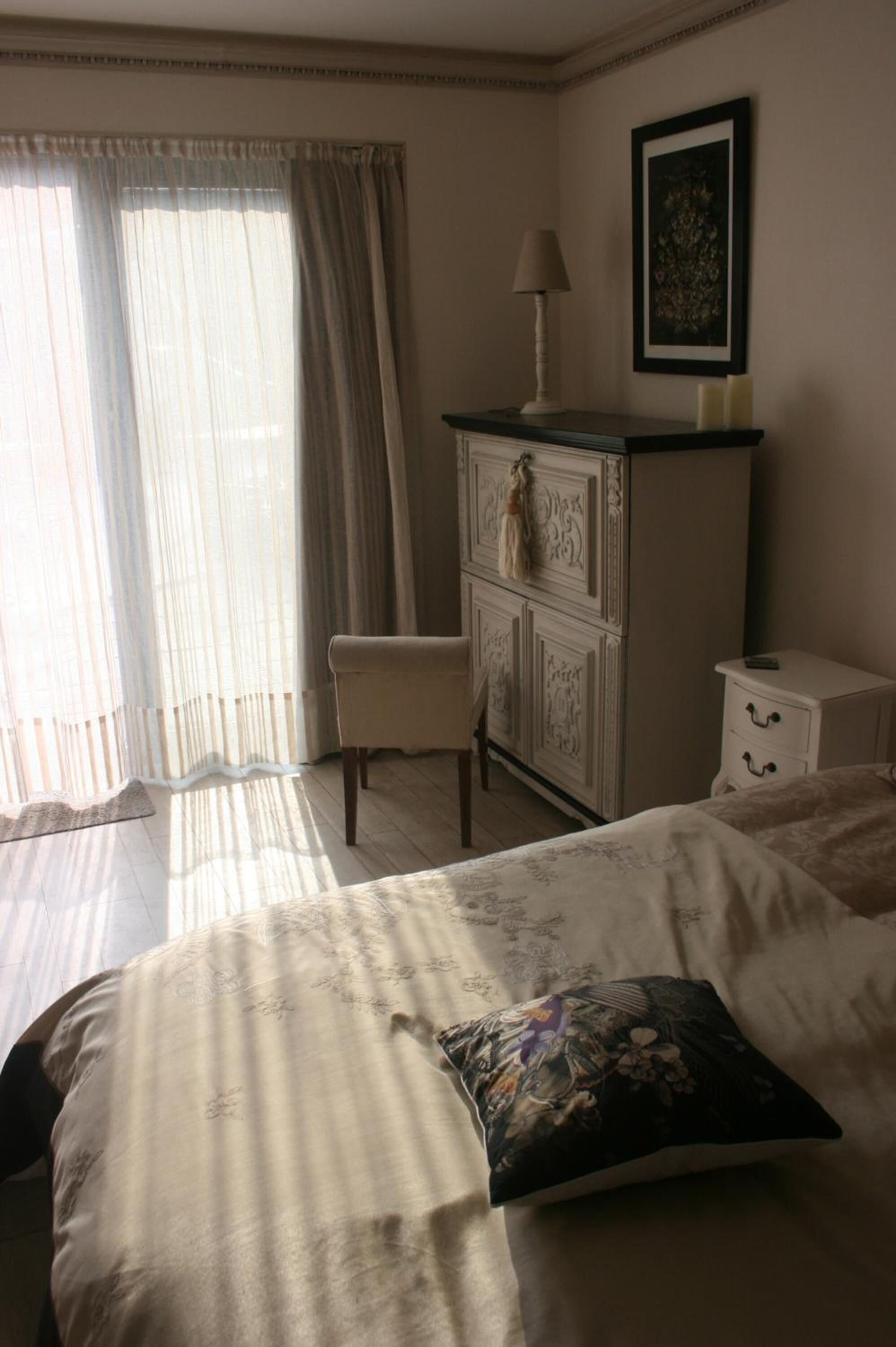 Un dormitorio con cama, una silla, cómoda, mesita de noche, lámpara y un cuadro en la pared.