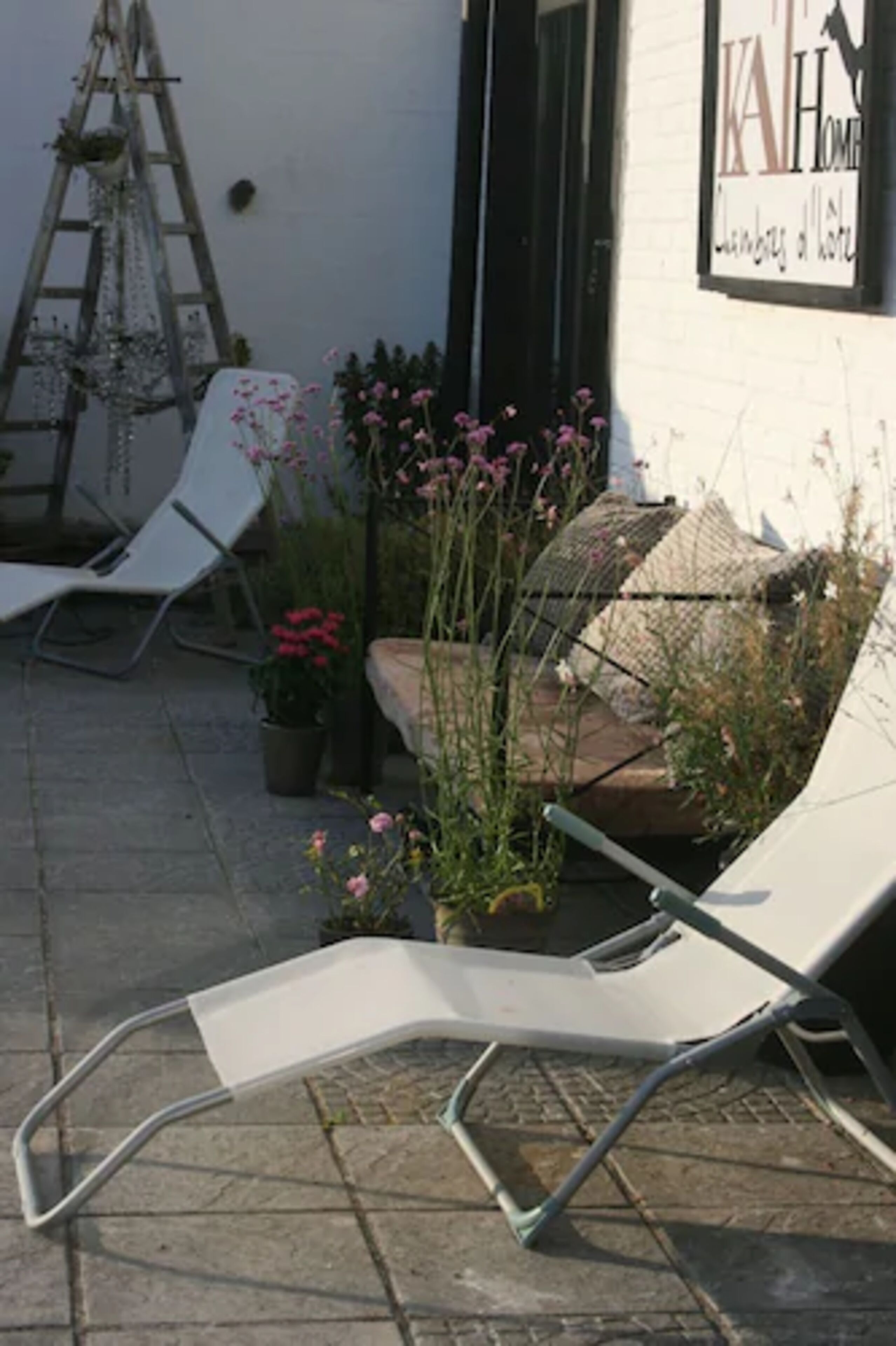 A patio with two white lounge chairs, a ladder, and potted plants with pink flowers.
