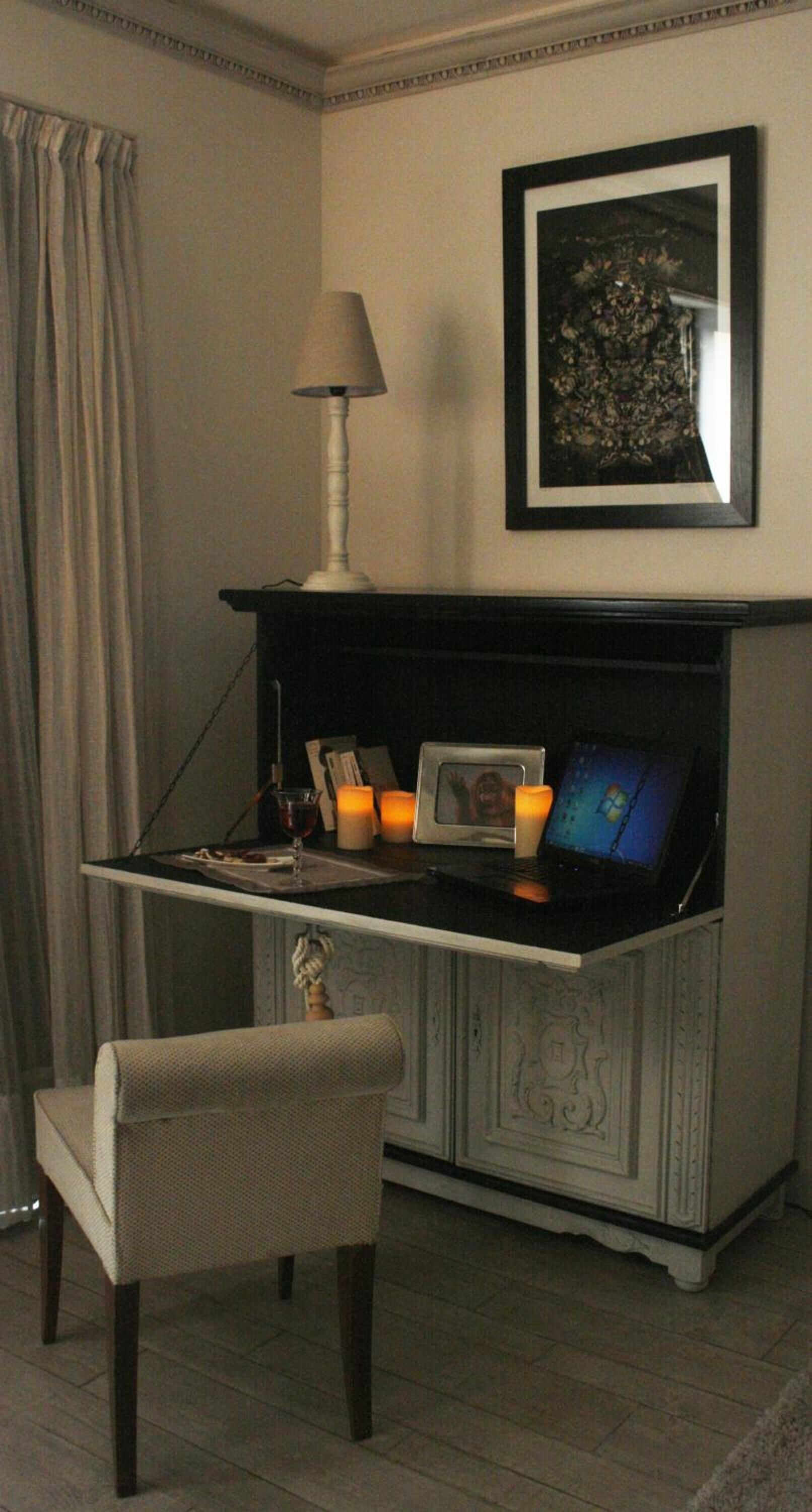 A corner room with a desk, chair, lamp, framed picture, and candles.