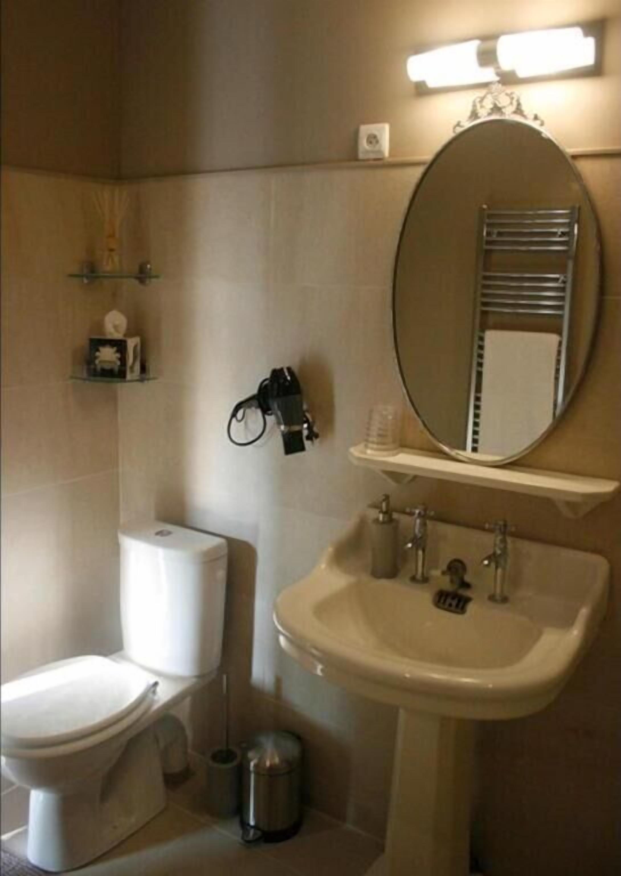 A bathroom with a toilet, sink, and mirror. There are shelves with toiletries and a trash can.