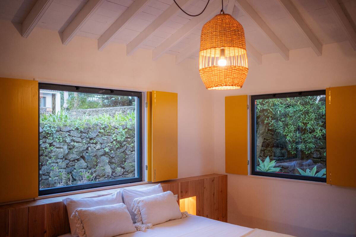Un dormitorio con una cama grande, cabecera de madera y dos ventanas con marcos amarillos.