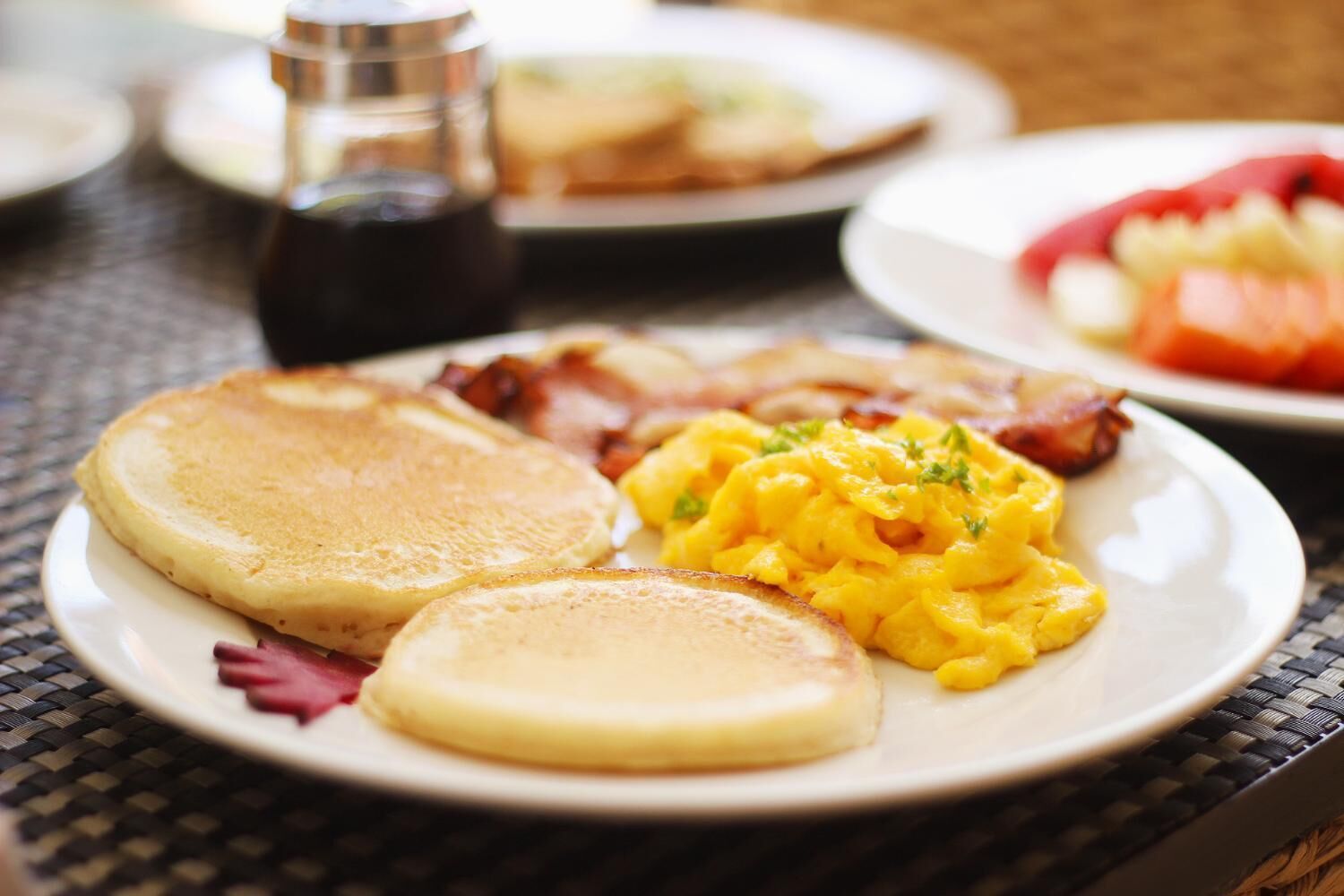 A breakfast plate with pancakes, scrambled eggs, and bacon on a table.