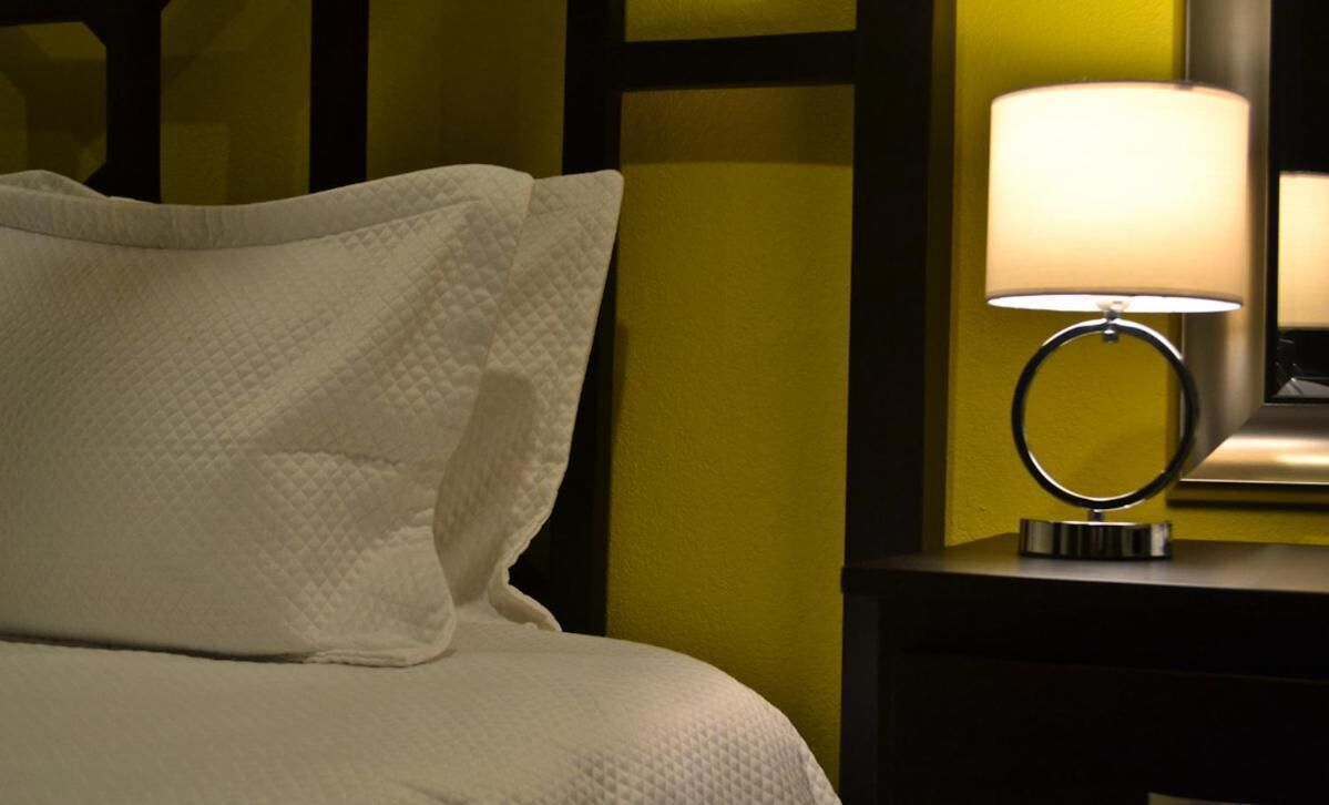 Una cama bien hecha con ropa blanca, una lámpara de noche y un mesita de noche oscura frente a una pared amarilla.
