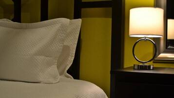 Una cama bien hecha con ropa blanca, una lámpara de noche y un mesita de noche oscura frente a una pared amarilla.