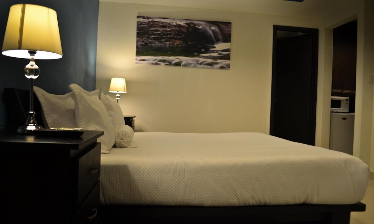 Ein ordentlich bezogenes Bett mit weißer Decke und Kissen, eine Nachttischlampe, ein an der Wand hängendes Bild von einem Wasserfall und eine Tür zu einem Küchenbereich mit einer Mikrowelle.