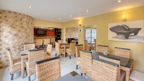 A dining area with wicker chairs and tables, a kitchen in the background, and a stone wall with a television. - Wayak Hotel & Suites (Managua)