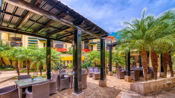 An outdoor patio with wicker furniture, palm trees, and a covered area with a metal roof. - Wayak Hotel & Suites (Managua)