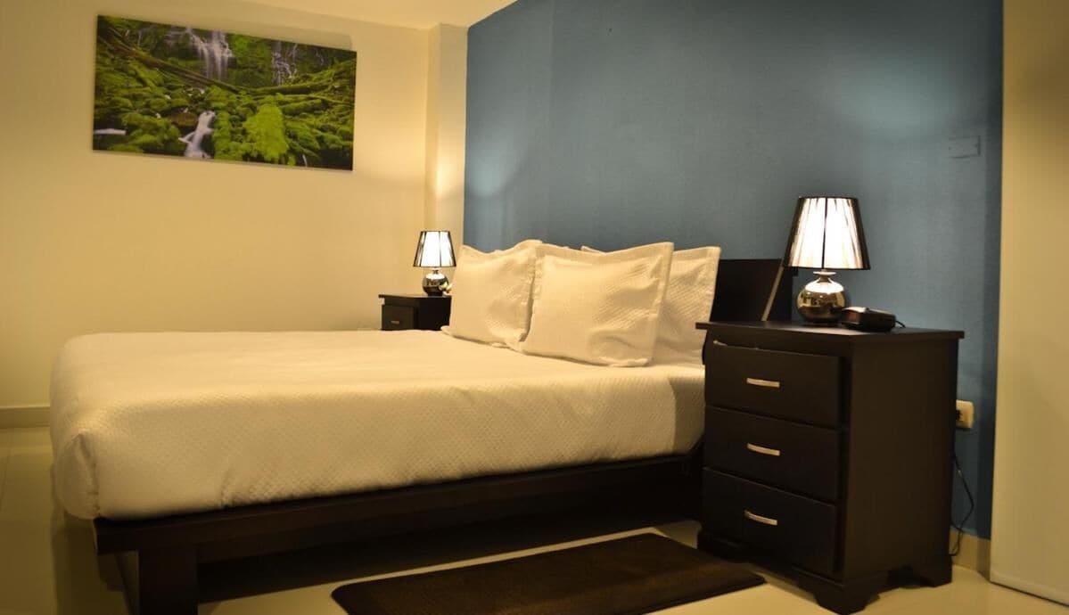 A bedroom with a bed, two bedside tables with lamps, a nightstand, a wall-mounted picture, and a dark-colored dresser.