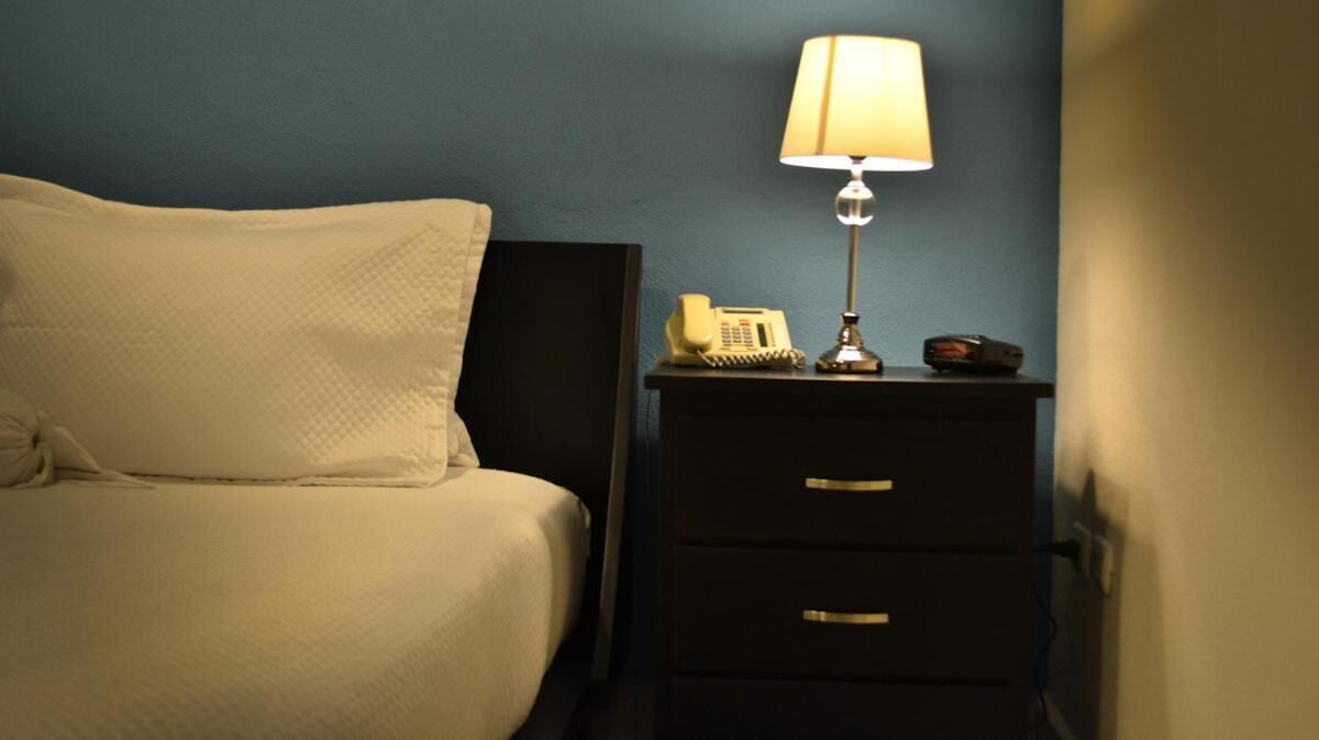 Un lit bien fait, avec une couette blanche et des oreillers, une table de chevet avec une lampe, un téléphone et un petit appareil électronique.