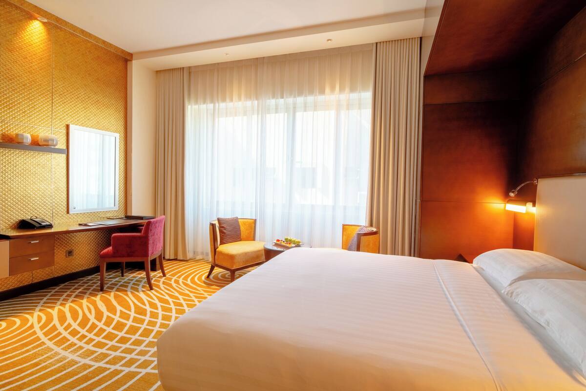 Ein Hotelzimmer mit einem großen Bett, einem Schreibtisch mit Stuhl, einem kleinen Tisch mit Früchten und einem großen Fenster mit Vorhängen.