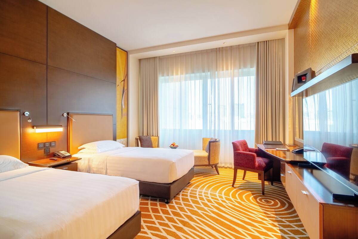Ein Hotelzimmer mit zwei Betten, einem Schreibtisch mit Computer, einem Stuhl und einem großen Fenster mit Vorhängen.