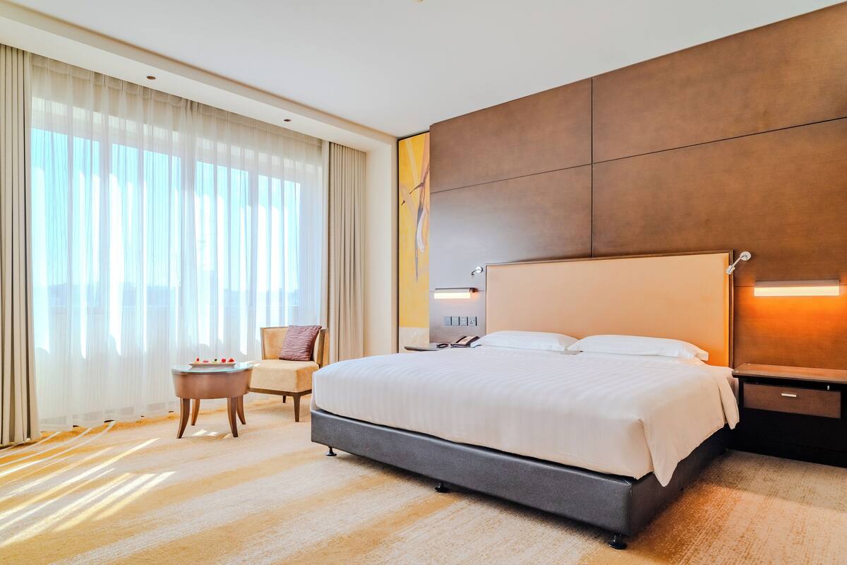 Ein Hotelzimmer mit einem großen Bett, einem Nachttisch, einem Sessel und einem Fenster mit Vorhängen.