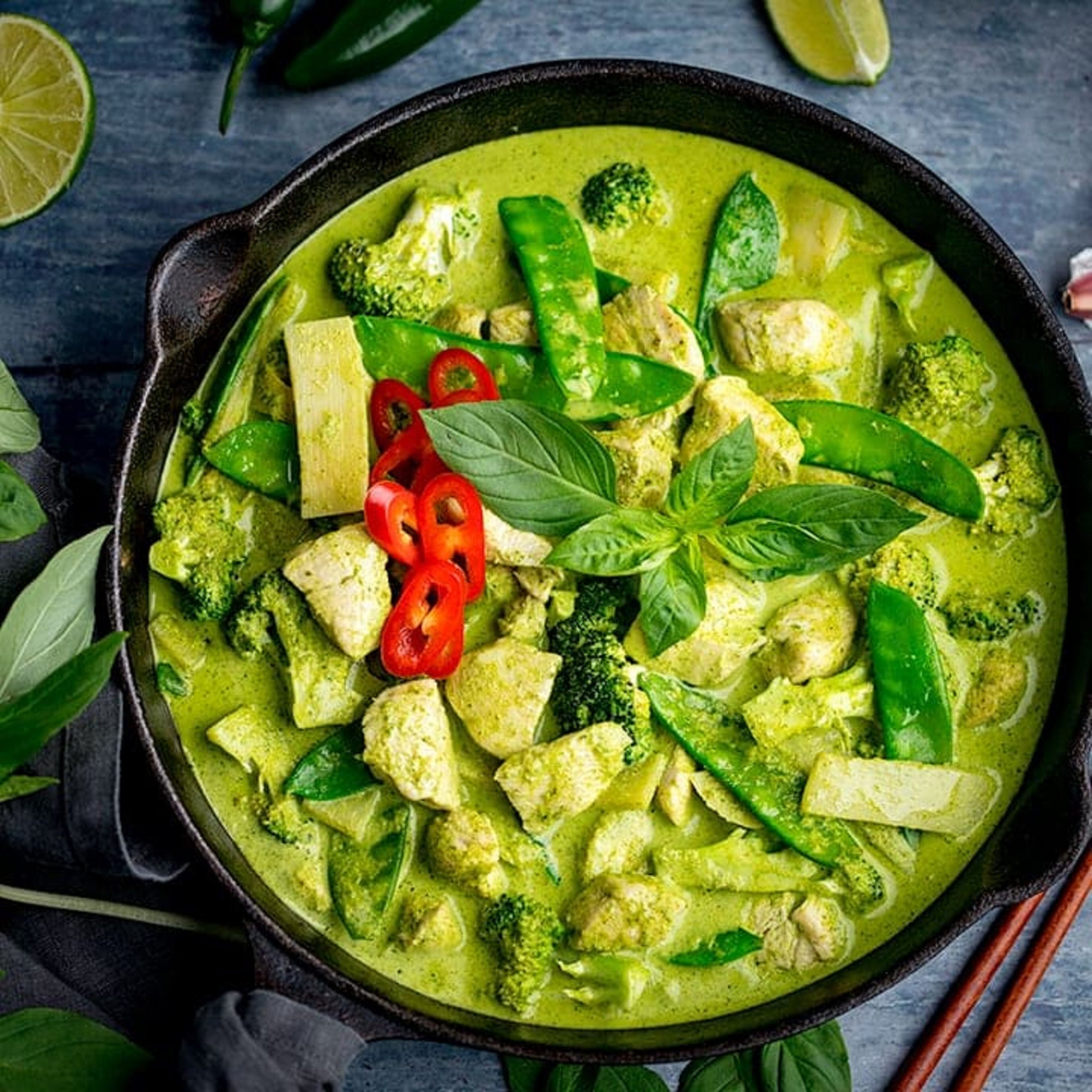 Een kom groene curry met groenten en kip, versierd met basilicumblaadjes en rode chilischijfjes.