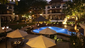 Un área de piscina con sombrillas y sillas de descanso, rodeada de edificios con balcones e iluminación exterior.