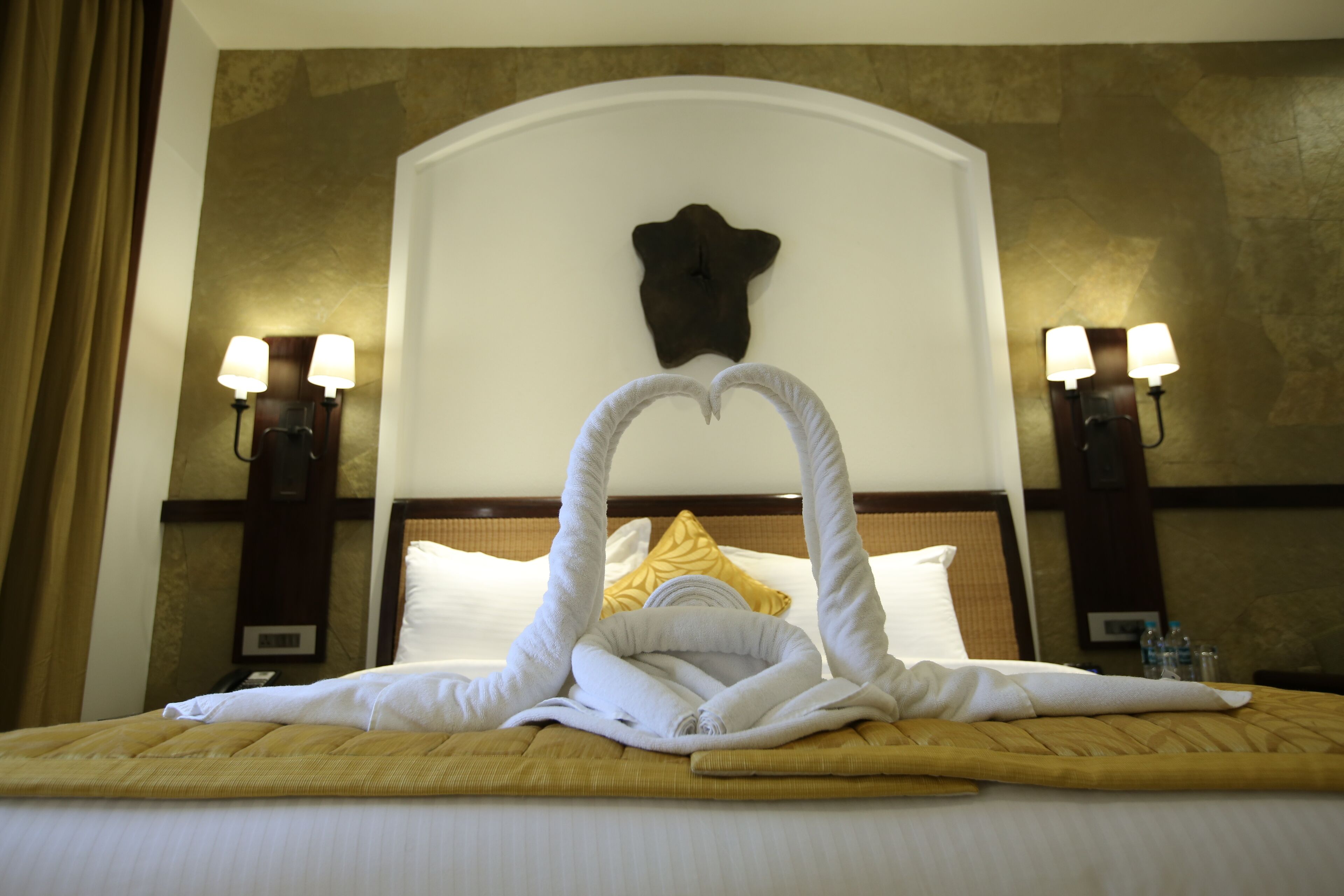 A hotel room with a bed, a heart-shaped towel arrangement, two wall-mounted lamps, and a decorative wall piece above the bed.