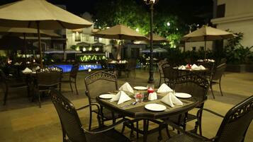 Un área de comedor al aire libre con mesas, sillas, sombrillas y una piscina al fondo.