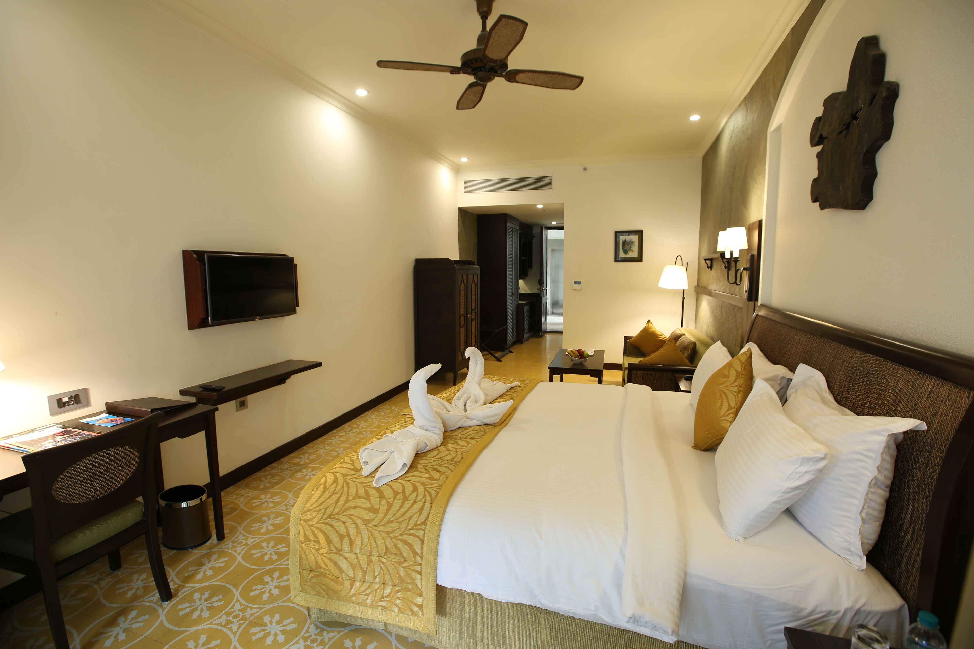A hotel room with a bed, a desk, a television, a ceiling fan, and a hallway.