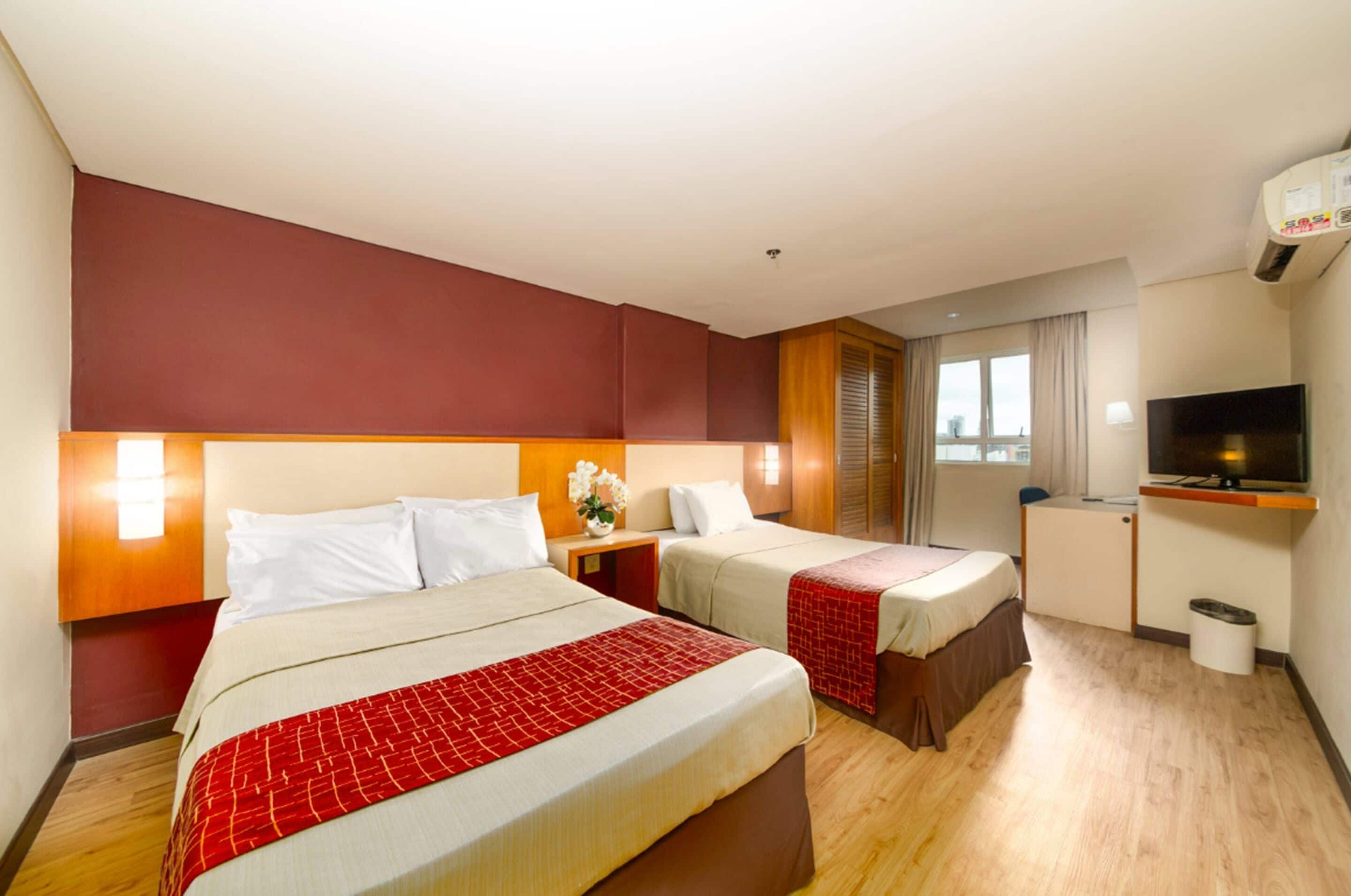 Quarto de hotel com duas camas, televisão, escrivaninha e janela com cortinas.