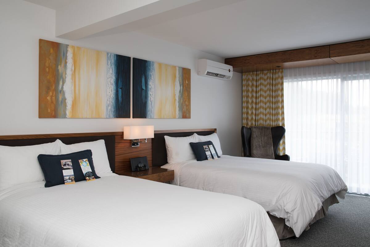 Habitación de hotel con dos camas, un cuadro abstracto grande, un equipo de aire acondicionado, una mesita de noche con lámpara y una ventana con cortinas translúcidas.