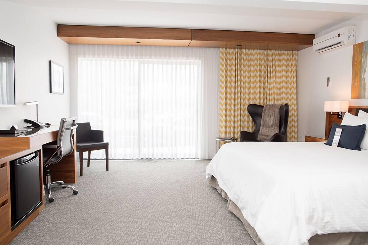 Una habitación de hotel con cama, escritorio, silla y unidad de aire acondicionado.