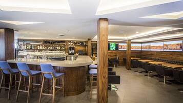 Eine moderne Bar mit einer zentralen Bar, verschiedenen Sitzmöglichkeiten inklusive Barhocker und Tische, und einem großen Bildschirm, der eine schöne Aussicht zeigt.