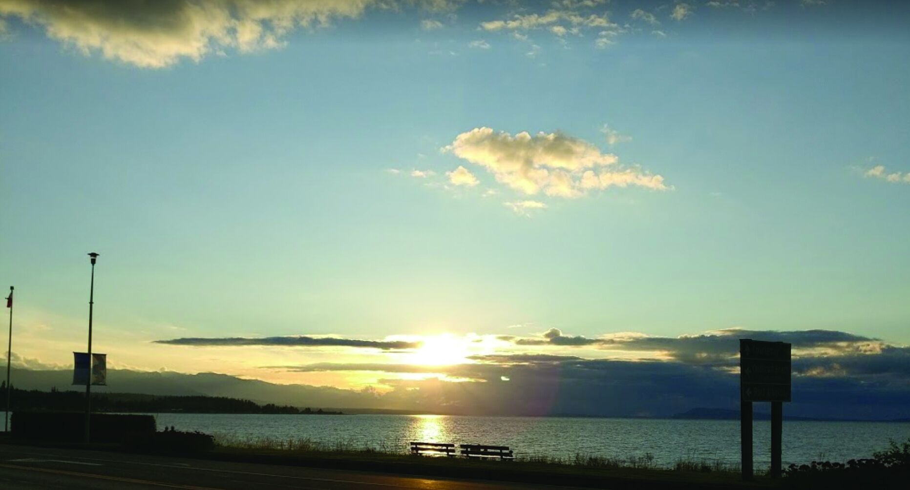 Auringonlasku vesistön yllä, etualalla penkki ja opastekyltti.