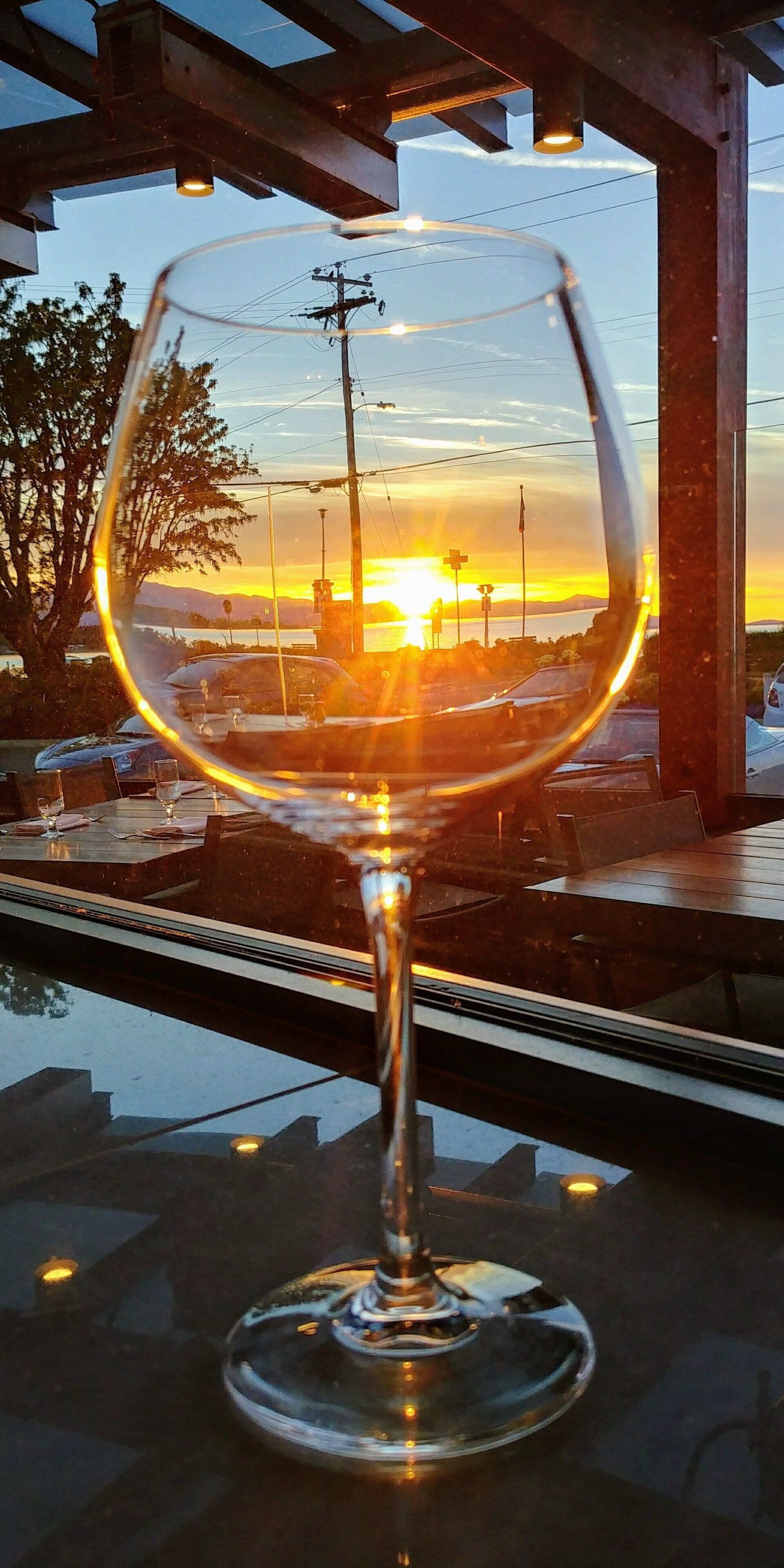 A glass of wine on a table with a view of a sunset over a body of water and mountains.