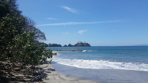Una playa con cielos despejados, aguas tranquilas y formaciones rocosas a lo lejos.