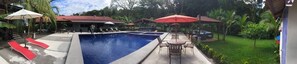 A swimming pool with lounge chairs and umbrellas, surrounded by greenery and a covered patio area. - Costa Del Sol (Tárcoles)