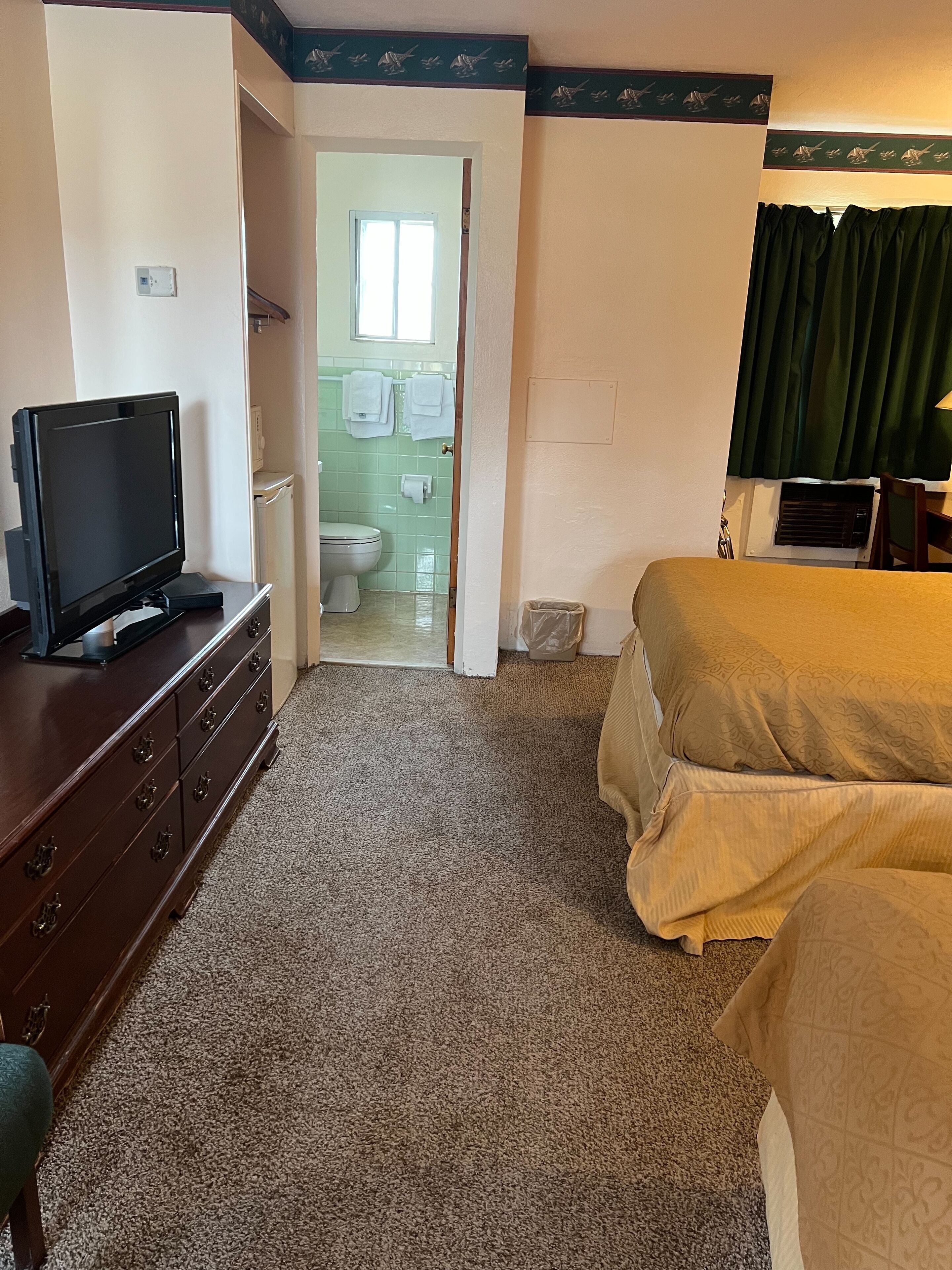 A hotel room with a television on a dresser, a bed with a beige cover, a carpeted floor, and a bathroom visible through an open door.