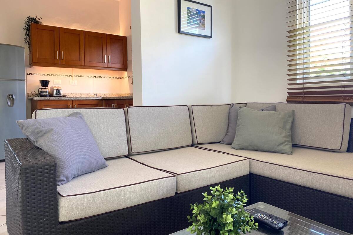 A modern living room with a sectional sofa, a coffee table with a remote control, and a kitchen in the background.