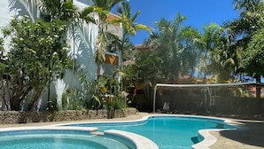 A swimming pool with a curved design, surrounded by lush greenery and a white building.