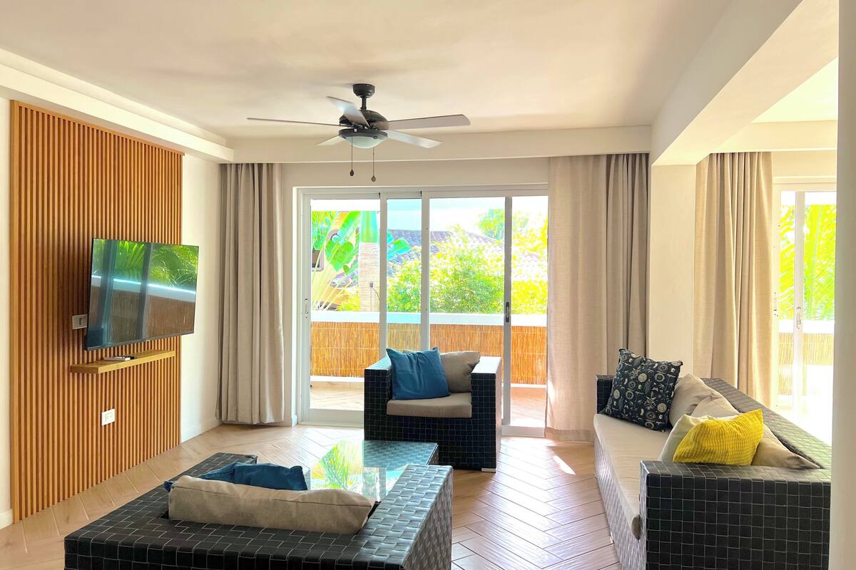 A modern living room with a flat-screen TV mounted on a wooden panel, a ceiling fan, and a sliding glass door leading to an outdoor area with a view of greenery.