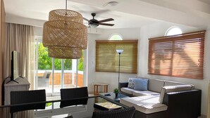 A modern living room with wicker furniture, a ceiling fan, and large windows with blinds.