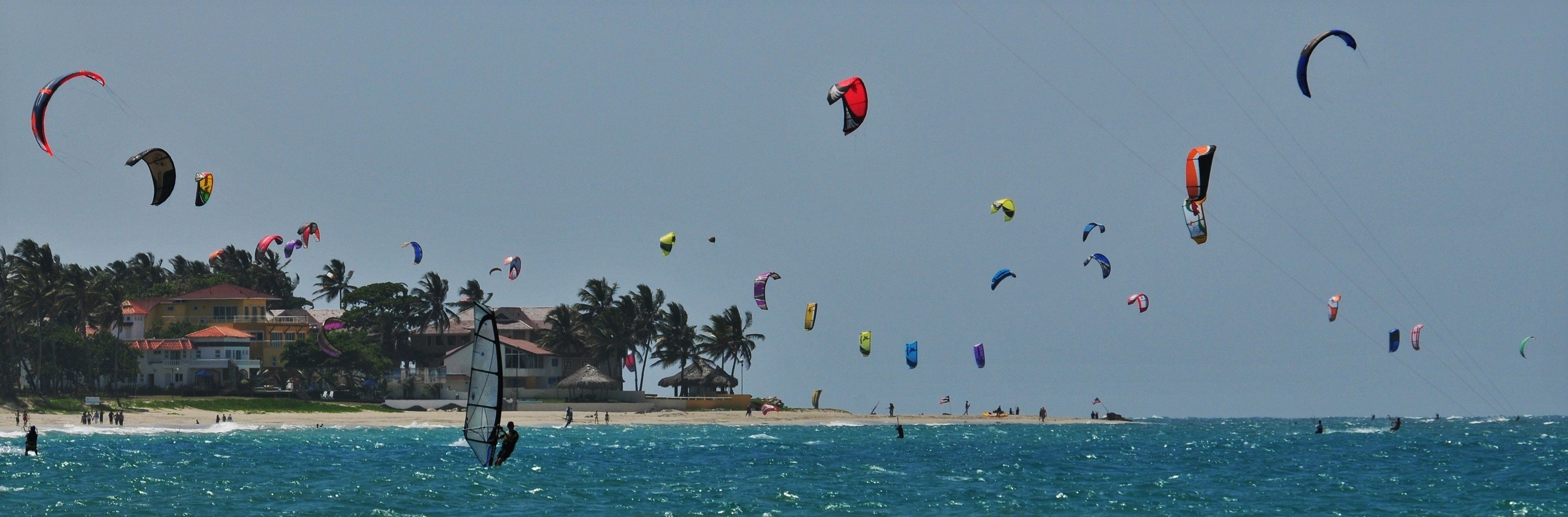 Una playa con varios cometas volando en el cielo, palmeras y un edificio con techo rojo al fondo.