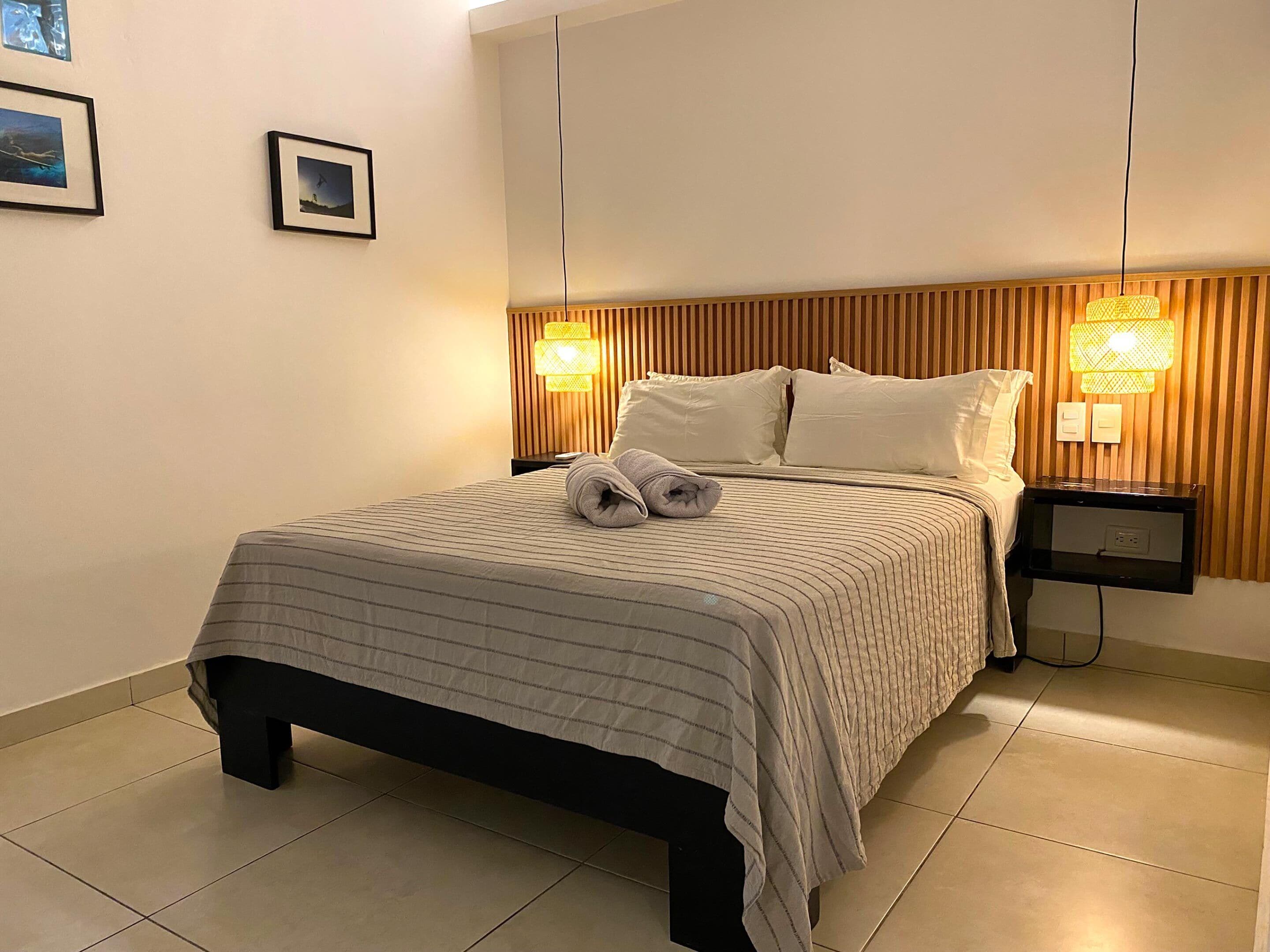 A neatly made bed with a striped bedspread, two pillows, and a rolled towel on the bed. There are two bedside tables with lamps, a wall-mounted shelf, and two framed pictures on the wall.