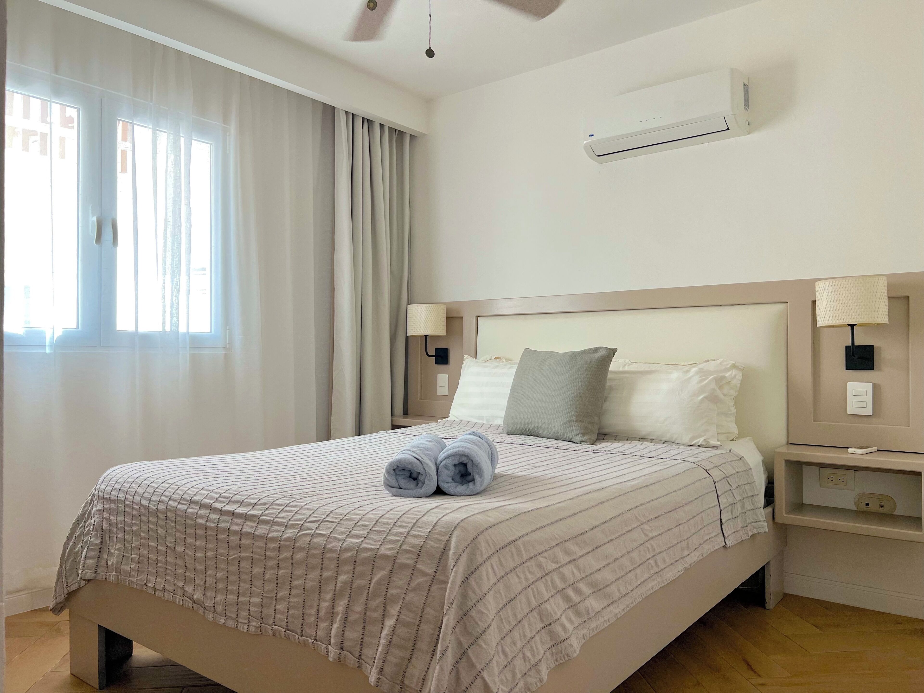 Una cama bien tendida con una colcha a rayas, dos toallas enrolladas y una almohada. Hay una mesita de noche con una lámpara, un aire acondicionado de pared y una ventana con cortinas translúcidas.