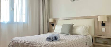 Una cama bien tendida con una colcha a rayas, dos toallas enrolladas y una almohada. Hay una mesita de noche con una lámpara, un aire acondicionado de pared y una ventana con cortinas translúcidas.